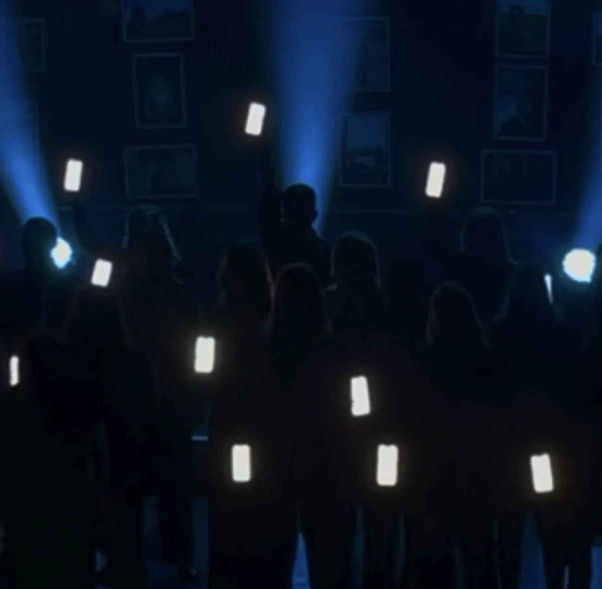 Performers silhouetted on a dark stage under blue beams, holding up phones whose screens glow white on cue